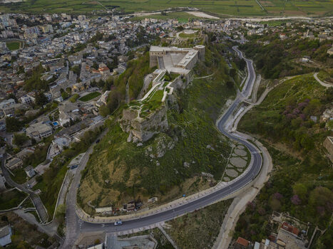 Aerial view of Gjirokaster Fortress perched on a rocky hill overlooking the historic city with winding roads and lush green surroundings in Gjirokaster Gjirokaster County Albania. Aerial view of Gjirokaster Fortress perched on a rocky hill overlooking the historic city with winding roads and lush green surroundings in Gjirokaster Gjirokaster County Albania.