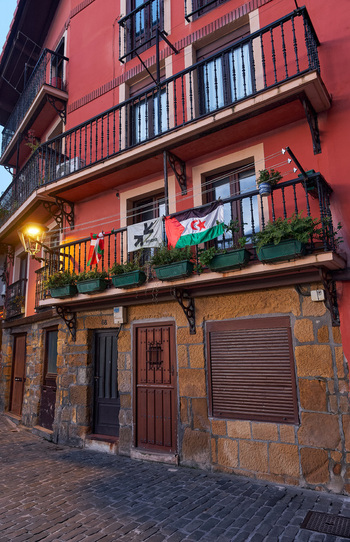 Pasai Donibane Spain 09 March 2026 View of the historic harbor street featuring a red building with balconies adorned with flags and flower boxes above a cobblestone path.