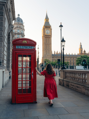View of a woman in a scarlet dress pausing by a vivid red telephone box with the timeless Big Ben rising majestically under a soft sky London England United Kingdom. View of a woman in a scarlet dress pausing by a vivid red telephone box with the timeless Big Ben rising majestically under a soft sky London England United Kingdom.