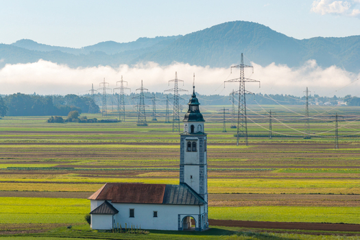 Aerial view of a historic church with a bell tower set amidst agricultural fields with power lines and mountains under a misty sky in Kranj Slovenia. Aerial view of a historic church with a bell tower set amidst agricultural fields with power lines and mountains under a misty sky in Kranj Slovenia.