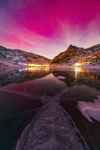 View of a frozen lake reflecting a vibrant pink night sky and snow covered mountains with village lights in Madesimo Lombardy Italy. View of a frozen lake reflecting a vibrant pink night sky and snow covered mountains with village lights in Madesimo Lombardy Italy.