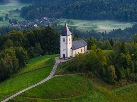 Aerial view of the Church of Saints Primus and Felician situated on a grassy hilltop surrounded by lush forests and rolling hills in Jamnik Kranj Slovenia.