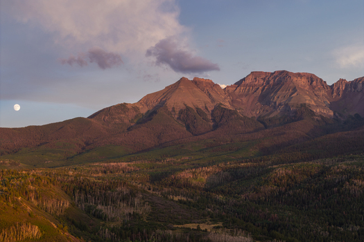 Aerial view of Mount Sneffels in the San Juan Mountain Range at golden hour with a visible moon over the rugged peaks and forested slopes in Ouray Colorado United States. Aerial view of Mount Sneffels in the San Juan Mountain Range at golden hour with a visible moon over the rugged peaks and forested slopes in Ouray Colorado United States.