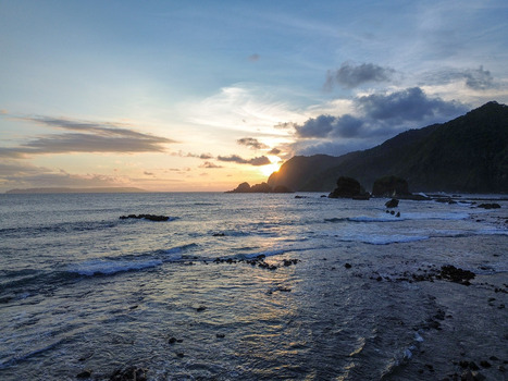 Aerial view of Papuma Beach at sunset with rocky outcrops gentle waves washing onto the shore and lush hills under a soft golden sky in Jember East Java Indonesia. Aerial view of Papuma Beach at sunset with rocky outcrops gentle waves washing onto the shore and lush hills under a soft golden sky in Jember East Java Indonesia.