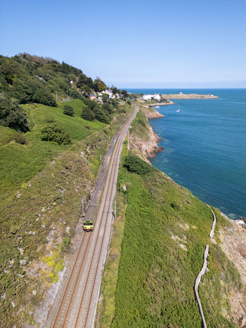 Aerial view of a train traveling along the coastal railway tracks in Dalkey surrounded by lush green hills and the blue sea in Dublin County Dublin Ireland. Aerial view of a train traveling along the coastal railway tracks in Dalkey surrounded by lush green hills and the blue sea in Dublin County Dublin Ireland.