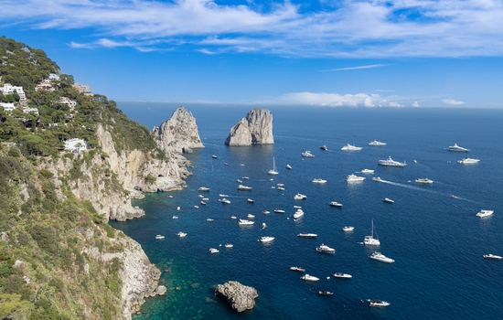 Aerial view of the iconic Faraglioni rock formations rising from the deep blue Tyrrhenian Sea with numerous yachts and boats along the rugged coastline of Capri Campania Italy. Aerial view of the iconic Faraglioni rock formations rising from the deep blue Tyrrhenian Sea with numerous yachts and boats along the rugged coastline of Capri Campania Italy.