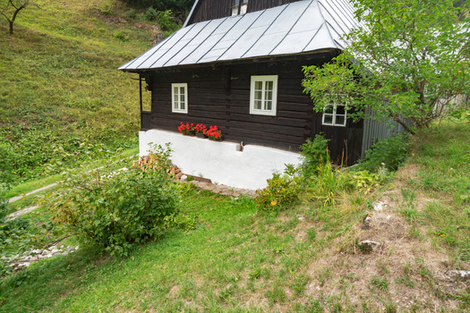 View of a charming dark wooden house with white trim and a silver roof sits nestled in a lush green hillside Prasnica Velka Fatra Slovakia. View of a charming dark wooden house with white trim and a silver roof sits nestled in a lush green hillside Prasnica Velka Fatra Slovakia.