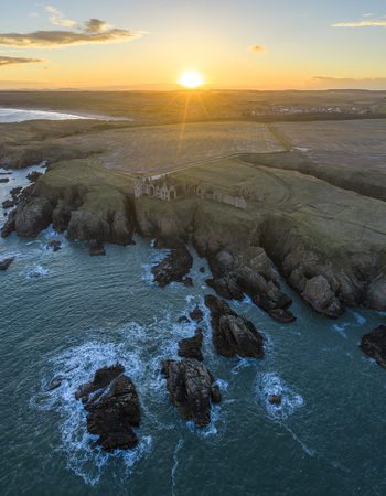Aerial view of Slains Castle ruins on a rugged cliff edge at sunset with golden light reflecting on the North Sea Cruden Bay Scotland United Kingdom. Aerial view of Slains Castle ruins on a rugged cliff edge at sunset with golden light reflecting on the North Sea Cruden Bay Scotland United Kingdom.