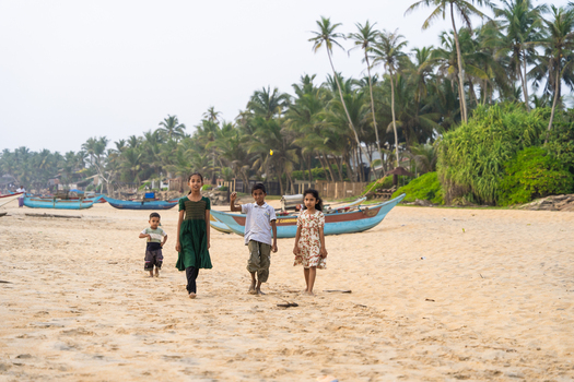 Sri Lanka 20 January 2026 View of four local children walking along a sandy beach with one boy waving traditional fishing boats and palm trees in the background.