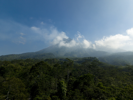 Aerial view of Mount Merapi volcano peak shrouded in clouds above a lush tropical forest under a clear blue sky in Kecamatan Cangkringan Daerah Istimewa Yogya Indonesia. Aerial view of Mount Merapi volcano peak shrouded in clouds above a lush tropical forest under a clear blue sky in Kecamatan Cangkringan Daerah Istimewa Yogya Indonesia.