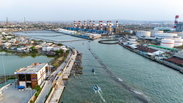 Aerial view of a power plant with tall chimneys and storage tanks situated along a water channel with small boats navigating the waterway in Semarang Central Java Indonesia. Aerial view of a power plant with tall chimneys and storage tanks situated along a water channel with small boats navigating the waterway in Semarang Central Java Indonesia.