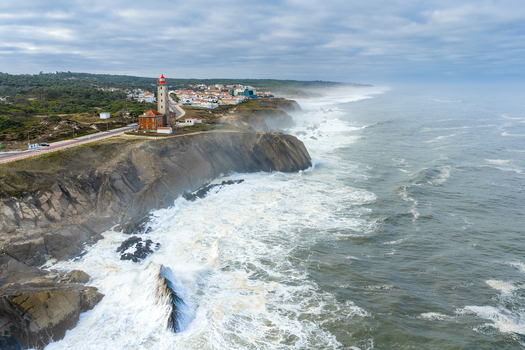 Aerial view of the red topped Farol Penedo da Saudade lighthouse on rugged cliffs with crashing waves and coastal town under a cloudy sky in Marinha Grande Leiria Portugal. Aerial view of the red topped Farol Penedo da Saudade lighthouse on rugged cliffs with crashing waves and coastal town under a cloudy sky in Marinha Grande Leiria Portugal.