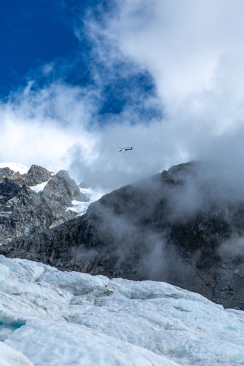 View of a helicopter flies above a glacier with jagged peaks piercing the clouds contrasting the icy blue expanse below Franz Josef Glacier West Coast Region New Zealand. View of a helicopter flies above a glacier with jagged peaks piercing the clouds contrasting the icy blue expanse below Franz Josef Glacier West Coast Region New Zealand.