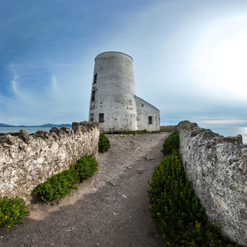 View of Twr Mawr Lighthouse at the end of a stone walled path with green bushes under a blue sky Isle of Anglesey Wales United Kingdom.