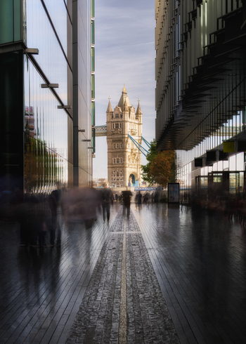 View of Tower Bridge framed by modern glass buildings with blurred people walking on a stone path in London Greater London United Kingdom.