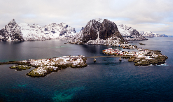 Aerial view of the Lofoten islands featuring snow covered mountains a bridge connecting small islands with red cabins and the dark blue sea in Nordland Norway.