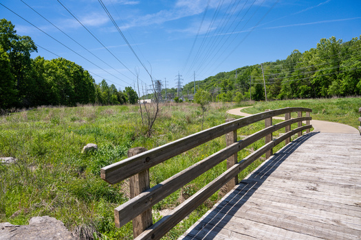 View of Greentree Park featuring a wooden bridge over a grassy field with power lines and lush trees under a clear blue sky Kirkwood Missouri United States. View of Greentree Park featuring a wooden bridge over a grassy field with power lines and lush trees under a clear blue sky Kirkwood Missouri United States.