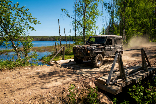 View of a black off road vehicle crossing a rustic wooden bridge on a dirt path beside the shores of Lake Ladoga in Sortavala Respublika Kareliya Russia.