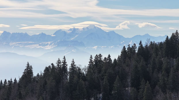 Aerial view of snow capped mountain peaks pierce through a hazy sky contrasting with the dark forest below Gex Auvergne Rhône Alpes France. Aerial view of snow capped mountain peaks pierce through a hazy sky contrasting with the dark forest below Gex Auvergne Rhône Alpes France.