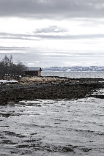View of a small wooden cabin on a rocky shoreline overlooking the cold sea with snow capped mountains under a cloudy sky Tromso Troms Norway.