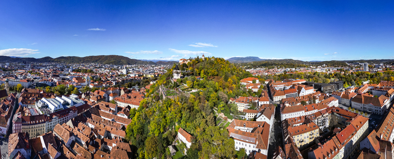 Aerial view of the vibrant red rooftops contrasting with the lush greenery of Schlossberg hill topped by the iconic clock tower Graz Styria Austria.