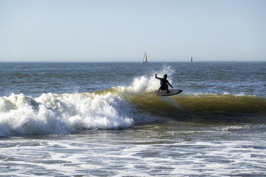 View of a surfer riding a breaking wave at Ereaga beach with white sea foam and sailboats on the horizon under a clear blue sky Algorta Euskadi Spain.