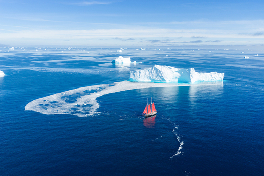 Aerial view of a red sailed ship sailing through deep blue arctic waters past a large white iceberg with scattered ice fragments under a clear sky Greenland. Aerial view of a red sailed ship sailing through deep blue arctic waters past a large white iceberg with scattered ice fragments under a clear sky Greenland.