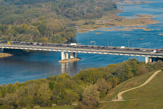 Aerial view of a modern bridge spanning the Vistula River with traffic flowing across the structure surrounded by lush green riverbanks and dense forest Warsaw Masovian Voivodeship Poland.