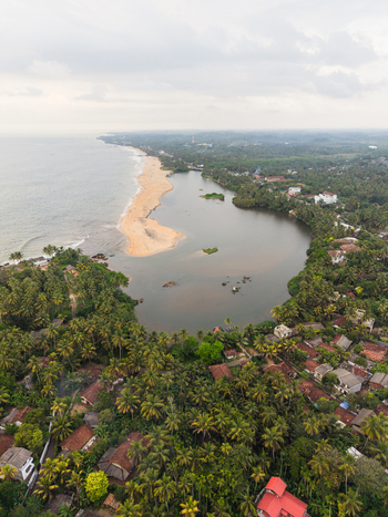 Aerial view of the tropical coastline where a lagoon meets the ocean surrounded by lush palm trees and residential rooftops under an overcast sky in Galle Southern Province Sri Lanka.
