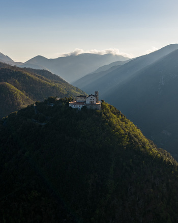 Aerial view of the Santuario del Santissimo Salvatore perched on a lush green hilltop surrounded by misty mountains and soft sunlight in Montella Campania Italy. Aerial view of the Santuario del Santissimo Salvatore perched on a lush green hilltop surrounded by misty mountains and soft sunlight in Montella Campania Italy.