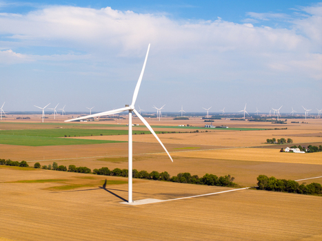 Aerial view of a wind turbine standing in a vast agricultural field with rows of turbines stretching across the horizon under a clear blue sky in Wolcott Indiana United States.