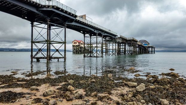 View of Mumbles Pier extending over the sea from a rocky shoreline under a dramatic overcast sky Mumbles Wales United Kingdom.