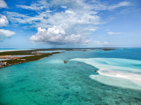Aerial view of the Man O War Cay Sandbar with boats anchored in turquoise water near the lush coastline under a cloudy sky in Rolletown Exuma The Bahamas. Aerial view of the Man O War Cay Sandbar with boats anchored in turquoise water near the lush coastline under a cloudy sky in Rolletown Exuma The Bahamas.