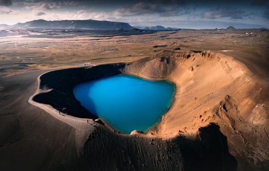 Aerial view of the turquoise Viti volcanic crater lake surrounded by rugged brown slopes and a vast barren landscape under a cloudy sky in Iceland.