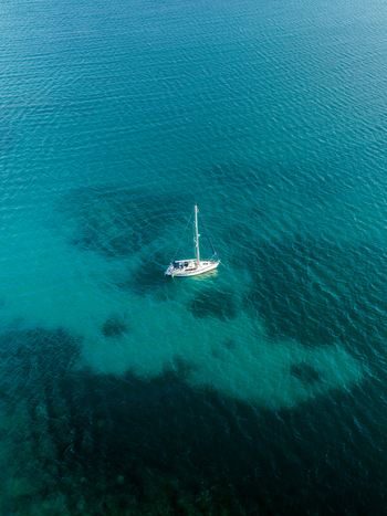 Aerial view of a lone white sailboat adrift on the shimmering turquoise waters casting a dark shadow on the seabed below Goro South Province New Caledonia. Aerial view of a lone white sailboat adrift on the shimmering turquoise waters casting a dark shadow on the seabed below Goro South Province New Caledonia.