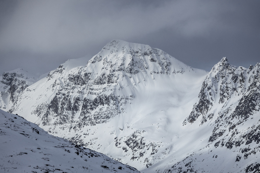 View of a steep snow covered mountain peak with rugged slopes under a dramatic cloudy sky in Ersfjord Troms Norway. View of a steep snow covered mountain peak with rugged slopes under a dramatic cloudy sky in Ersfjord Troms Norway.