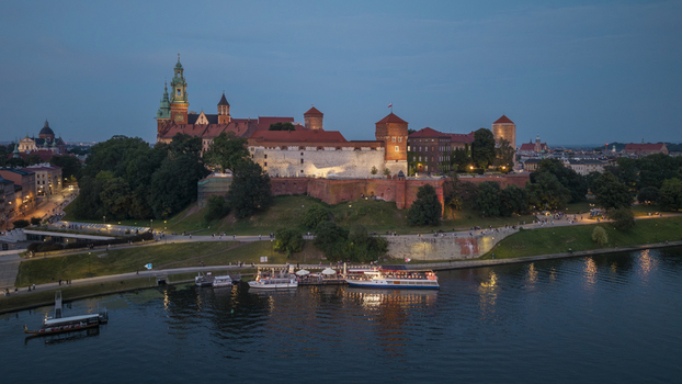 Aerial view of Wawel Castle illuminated at dusk overlooking the Vistula River with boats docked along the embankment and people walking nearby in Krakow Lesser Poland Voivode Poland. Aerial view of Wawel Castle illuminated at dusk overlooking the Vistula River with boats docked along the embankment and people walking nearby in Krakow Lesser Poland Voivode Poland.