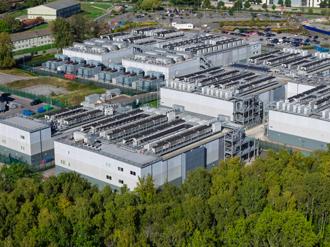 Aerial view of a large data center complex featuring multiple industrial buildings rows of rooftop cooling units and surrounding green trees United Kingdom.