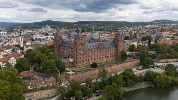 Aerial view of Schloss Johannisburg a historic red sandstone castle surrounded by lush trees and the Main river flowing nearby under a cloudy sky Aschaffenburg Bayern Germany.