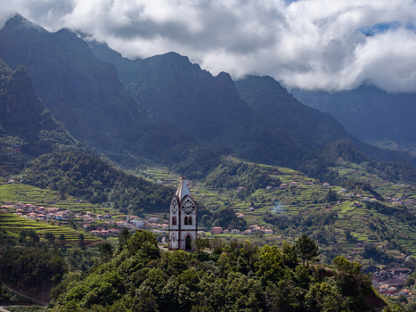 Aerial view of a chapel tower standing on a lush green hill surrounded by terraced fields and dramatic mountains in Madeira. Aerial view of a chapel tower standing on a lush green hill surrounded by terraced fields and dramatic mountains in Madeira.