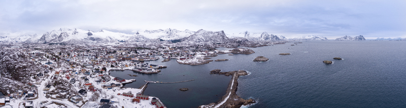 Aerial view of Lofoten islands featuring a snowy coastal town with colorful houses a harbor and dramatic snow capped mountains under a cloudy sky in Nordland Norway.