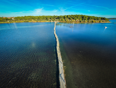 Aerial view of a narrow causeway stretching across shimmering waters towards a verdant island a contrast of blue and green under a clear sky Premantura Istria County Croatia.