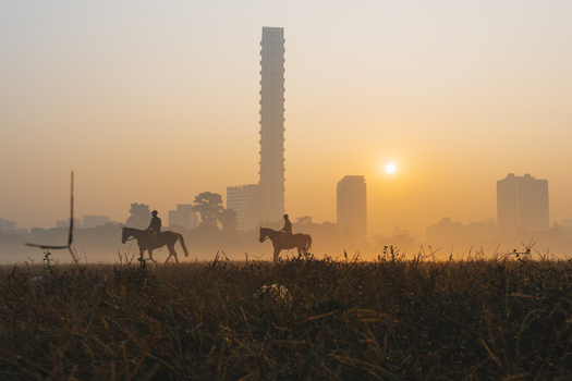 View of silhouetted riders astride horses traversing a sun kissed field with the iconic towering 42 building piercing through the misty Kolkata skyline Kolkata West Bengal India. View of silhouetted riders astride horses traversing a sun kissed field with the iconic towering 42 building piercing through the misty Kolkata skyline Kolkata West Bengal India.