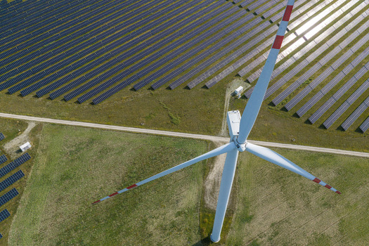 Aerial view of a large wind turbine with red tipped blades standing next to extensive rows of solar panels in a grassy field in Poland.