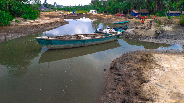 Aerial view of a wooden boat moored in a narrow waterway in Bukuma with people standing on the muddy bank near small huts and lush tropical vegetation in Buguma Rivers Nigeria.