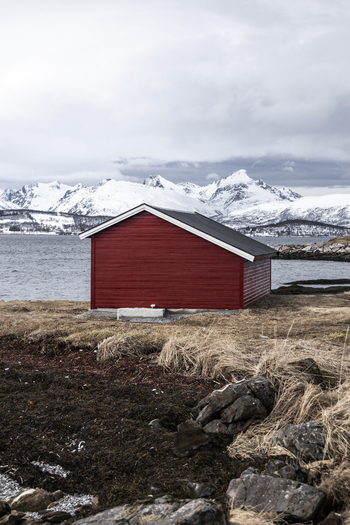 View of a traditional red wooden cabin on the shore with snow capped mountains across the fjord under an overcast sky Tromso Troms Norway.
