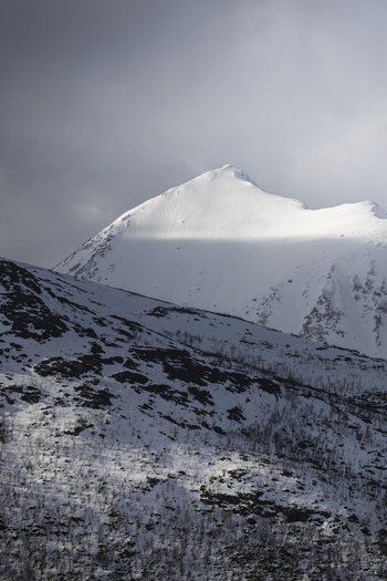 View of a snow covered mountain peak with rocky slopes and sunlight hitting the summit under a dark dramatic sky Kvaloya Troms Norway. View of a snow covered mountain peak with rocky slopes and sunlight hitting the summit under a dark dramatic sky Kvaloya Troms Norway.