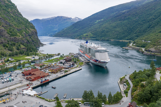 Aerial view of a large cruise ship docked at the port surrounded by steep green mountains calm fjord waters and traditional wooden buildings in Flam Vestland Norway. Aerial view of a large cruise ship docked at the port surrounded by steep green mountains calm fjord waters and traditional wooden buildings in Flam Vestland Norway.