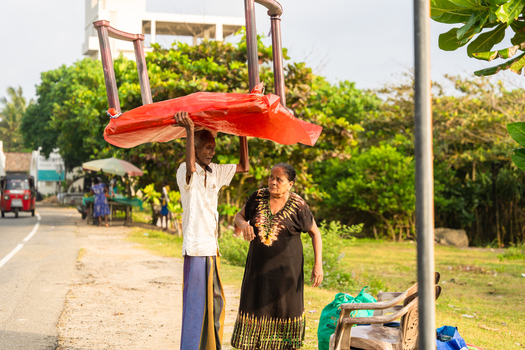 Sri Lanka 15 January 2026 View of an elderly man carrying a large chair on his head covered with red plastic while walking along a roadside with a woman.