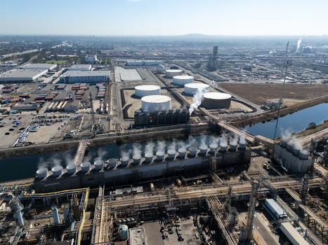 Aerial view of an industrial oil refinery complex with large storage tanks and cooling towers emitting steam over a canal in Carson California United States.
